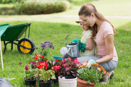 In the foreground of the garden, a young woman and a child are kneeling on the grass, engaging in gardening activities. They are surrounded by a variety of potted flowers and plants, including red, purple, and orange blooms, with some in decorative pots and others in containers. The woman is wearing a light-colored t-shirt and jeans, while the child is dressed in a pale yellow shirt. They are using small gardening tools and a watering can, focusing on tending to their garden plants. To the left, there is a green and yellow wheelbarrow positioned on the neatly trimmed lawn, which features lush, dense grass typical of well-maintained outdoor spaces. In the background, a hedge borders the garden, and a paved pathway is partially visible, suggesting a structured layout typical for suburban gardens. The scene is outdoors during daylight hours, with natural light illuminating the vibrant colours of the flowers and the verdant greenery, consistent with a suburban garden possibly in Peckham, indicative of local gardening services that specialise in planting and outdoor maintenance. The overall setting is tranquil, well-kept, and showcases a small, productive garden environment suitable for family gardening activities.