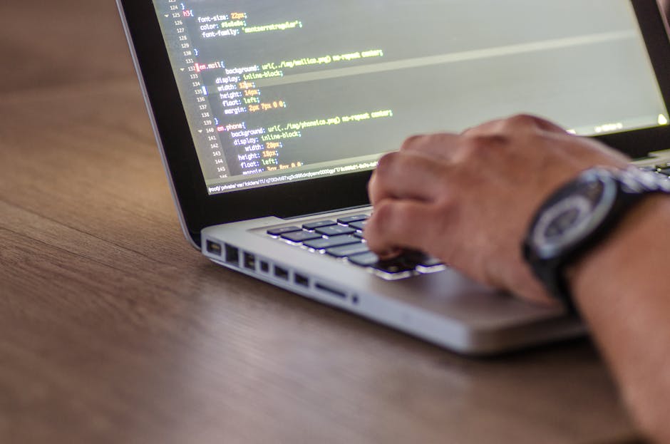 A close-up of a personâ€™s hands typing on a silver laptop with a black keyboard against a dark, blurred background. The laptop screen displays a dark-themed coding interface with lines of colorful co